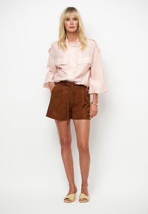 Lichtroze overhemd met knopen en twee borstzakken; bruine suède korte broek met ceintuur; gecombineerd met beige instapschoenen.