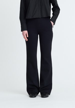 Person trägt schwarze ausgestellte Hose, schwarze Schuhe und ein schwarzes Langarmshirt, eine Hand in der Tasche und die andere an der Seite.