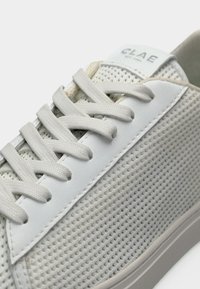 Λευκό αθλητικό παπούτσι με υφασμένες πλαϊνές επιφάνειες, λευκά κορδόνια και ετικέτα στη γλώσσα με την επιγραφή "CLAE" σε σκούρα γράμματα.