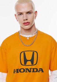 Orange bomulds T-shirt med et stort sort Honda-logo. Modellen bærer lag på lag af sølvkæder og en lille vedhæng.
