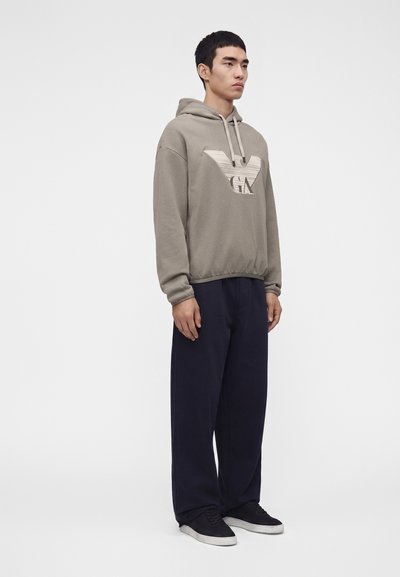 Homme debout portant un sweat à capuche taupe avec un grand logo, un pantalon large bleu marine et des baskets noires sur un fond blanc uni.