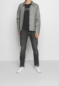Camisa a cuadros grises sobre una camiseta negra, combinada con jeans gris oscuro y zapatillas blancas. Tejido suave, cierre de botones, corte casual.