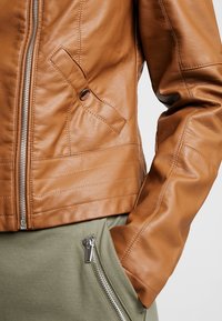 Hnědá kožená bunda s přiléhavým designem, s předním zipem, boční kapsou a prošívanými detaily podél lemu a rukávů.