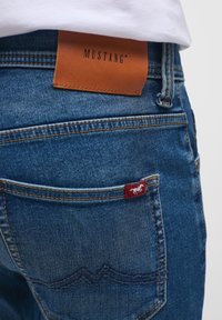 Jeans de denim azul con una etiqueta de cuero marrón marcada "MUSTANG", que presenta una etiqueta de tela rojo oscuro con un logo de caballo en el bolsillo trasero.