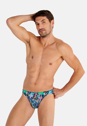 Fitte man poseert met één hand achter het hoofd en de andere op de heup, gekleed in kleurrijke zwembroek met patroon, tegen een effen achtergrond.