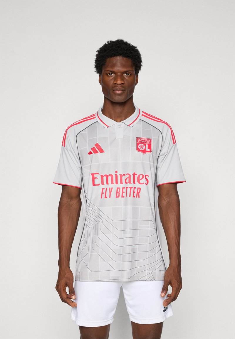 Homme portant un maillot de football Olympique Lyonnais gris avec des accents rouges et un short blanc, se tenant contre un fond blanc uni.