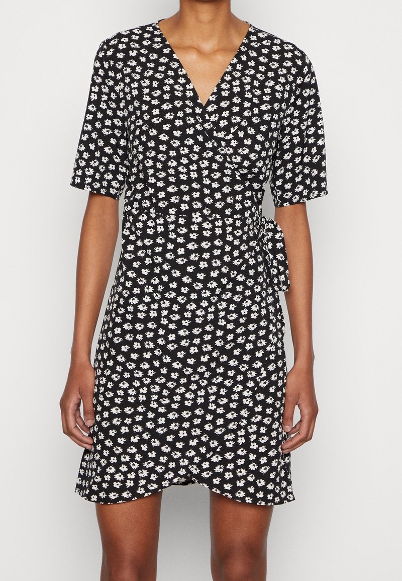 Femme portant une robe portefeuille noire à manches courtes avec un motif floral blanc, nouée à la taille, debout devant un fond uni.