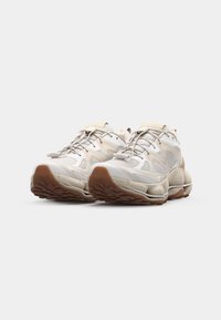 Light-colored sneakers with a textured mesh upper, contrasting rubber sole, and rounded, cushioned design. Laces are prominently featured.