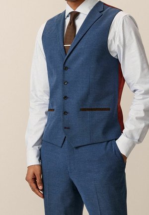 Homme portant un gilet et un pantalon bleu sur mesure avec une chemise blanche rayée et une cravate marron à pois, main dans la poche sur fond neutre.