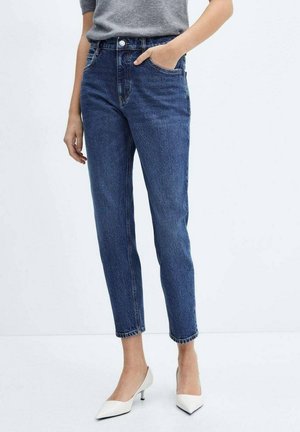 Vrouw draagt hoge, taps toelopende blauwe jeans met zakken aan de voorkant, gecombineerd met een ingestopte grijze blouse en witte kitten heels met een puntige neus.