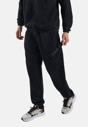 Man die zwarte Reebok trainingsbroek en zwart-witte Reebok sneakers draagt, staat met één been iets naar voren.