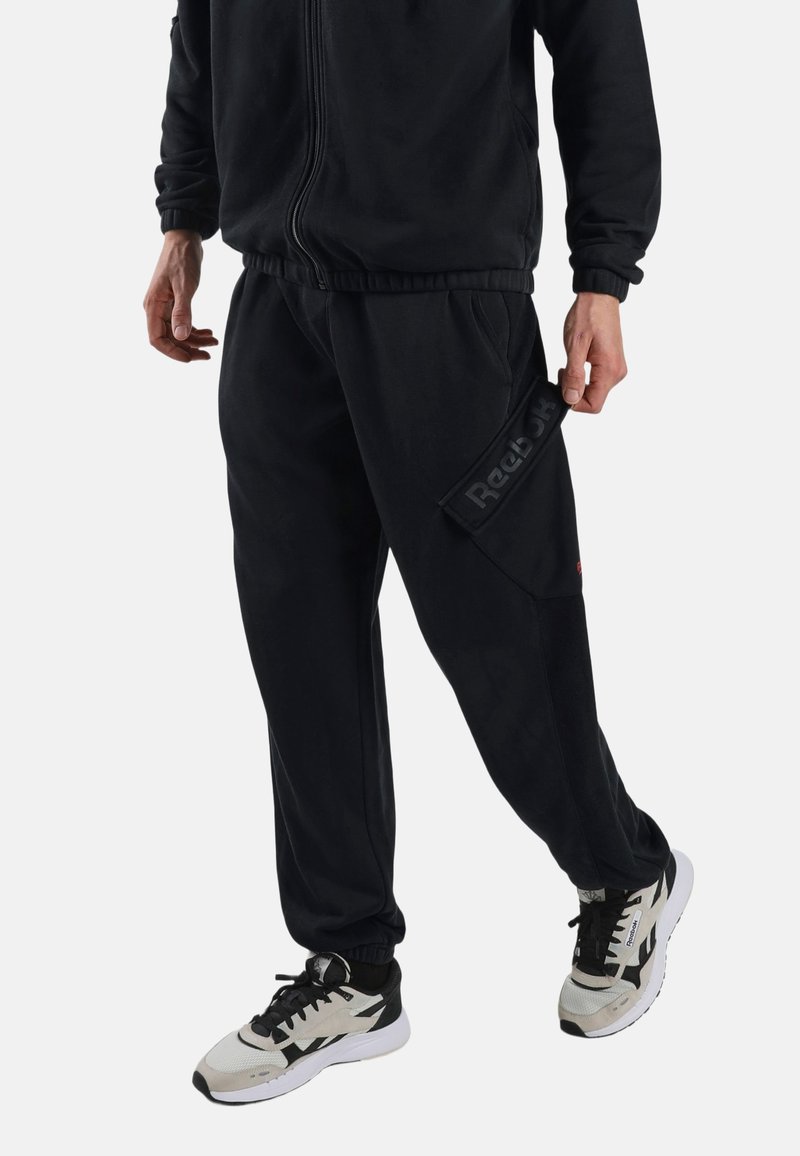 Man wearing black Reebok track pants and black-and-white Reebok sneakers, standing with one leg slightly forward.