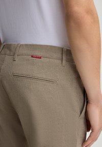 Pantaloni beige in tessuto testurizzato, dotati di una tasca posteriore e di una piccola etichetta rossa con la scritta "JOOP! JEANS." Design semplice e sartoriale.
