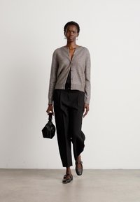 Lys grå teksturert cardigan, svart topp, svarte vide bukser, og svarte ankelstroppsko. Holder en liten svart håndveske. Nøytral bakgrunn.