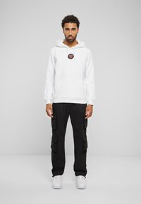 Hvit bomull hettegenser med frontlomme og logo, sammen med svarte cargo-bukser med flere sidelommer, og hvite joggesko.