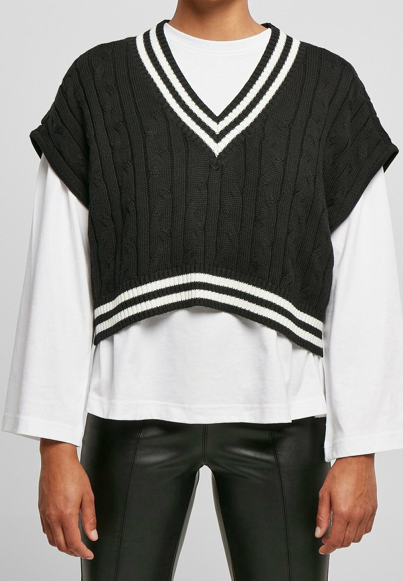 Schwarzer gestrickter, kürzerer Pullover mit V-Ausschnitt, der weiße Streifenakzente am Ausschnitt und Saum aufweist. Über einem weißen Langarmshirt getragen.