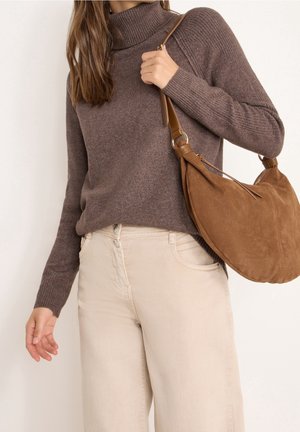 Femme portant un pull à col roulé marron et un pantalon beige clair, portant un grand sac à épaule en daim marron sur son épaule droite.