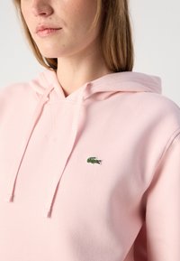 Hanorac roz cu o textură specială, având o buzunar tip kenguru, glugă cu șnur și un mic logo verde de crocodil pe piept.