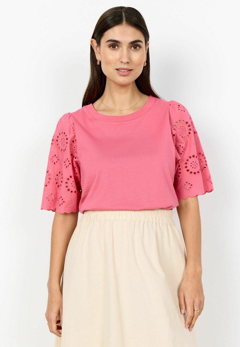 Kvinde med langt mørkt hår iført en lyserød top med hulmønstrede pufærmer og et cremelangt, højtaljet skørt, stående foran en hvid baggrund.