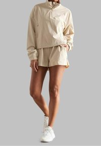 Polo beige léger avec une demi-fermeture éclair et des poignets élastiques, associé à un short assorti. Texture lisse avec une coupe décontractée. Chaussures de sport blanches.