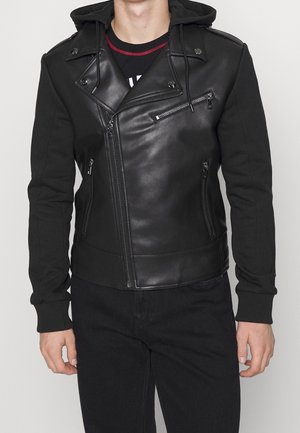 Mand iført sort asymmetrisk læderjakke med lynlåslommer foran, lynlåsdetaljer og fastmonteret sort hætte over sorte bukser.