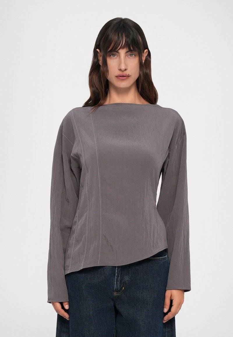 Maglia grigia a maniche lunghe con scollatura a barca, caratterizzata da dettagli sulle cuciture laterali e un orlo leggermente curvo, abbinata a jeans in denim scuro.