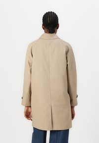 Manteau beige avec un col pointu, des manches raglan et une fente centrale au dos. Texture lisse avec des accents de boutons sur les poignets.