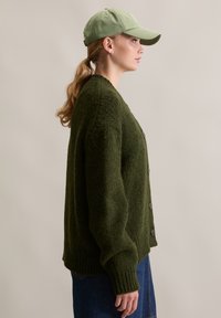 Cardigan en tricot vert olive à coupe ample, avec de gros boutons et des poignets côtelés, associé à une casquette vert clair en profil latéral.