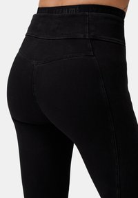 Primo piano di una persona che indossa leggings neri a vita alta con cuciture visibili e una cintura parzialmente visibile con del testo.