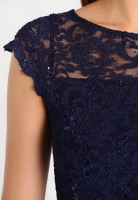 Marinblå spetsklänning med blommönster, genomskinliga axelbandsärmar och paljetterade detaljer. Texturerat tyg med intrikata detaljer.