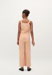 Lys rosa jumpsuit med et tekstureret, abstrakt mønster. Firkantet rygudskæring, brede ben og tynde stropper. Båret med sorte sko.