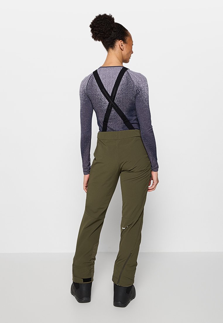 Frau mit lockigem Haar im Dutt trägt ein lila Langarmoberteil, olivgrüne Hosen mit schwarzen Hosenträgern und schwarze Stiefel und steht mit dem Rücken zur Kamera.