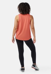 Mouwloze koraalkleurige tanktop met een losse fit en een gebogen zoom, gecombineerd met zwarte leggings. Gemaakt met sportieve schoenen met een grijs en oranje ontwerp.