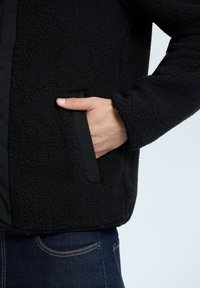 Mustassa fleece-takissa on teksturoitu ulkopinta, sileä sisävuori ja toinen sivutasku, joka on reunustettu kontrastoivalla mustalla trimmausnauhalla.