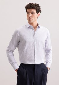 Uomo dai capelli ricci che indossa una camicia bianca a strisce verticali sottili e pantaloni scuri plissettati, con le mani nelle tasche, che guarda di lato.