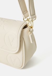 Beige leren tas met textuur en gouden hardware, voorzien van een gegraveerd logo en een afneembare schouderband. Compact rechthoekig van vorm met een klep sluiting.