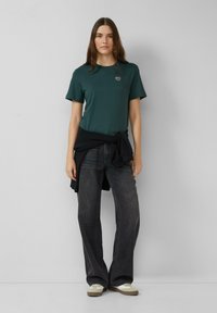 Donkergroene katoenen t-shirt met korte mouwen en een klein logo op de borst, gecombineerd met high-waisted zwarte spijkerbroek en witte sneakers.
