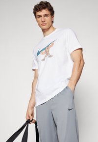 Hvid bomulds t-shirt med et farverigt tegneserie-grafik af en karakter med en baseballbat, parret med grå Nike bukser og sorte stropper.