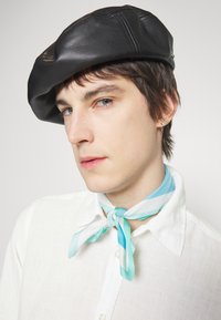 Beret de cuero negro con una textura suave, combinado con una bufanda de seda blanca y azul claro con un patrón, anudada al cuello, sobre una camisa de lino blanca.