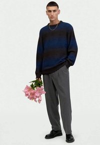 Knit sweater with blue and black stripes, loose fit, ribbed texture. Gray trousers, holding pink lilies. Black shoes. Simple gold chain necklace.