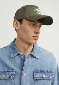 Gorra verde oliva con logotipo bordado en la parte frontal, combinada con una chaqueta de mezclilla azul clara con cuello abotonado y bolsillos en el pecho.