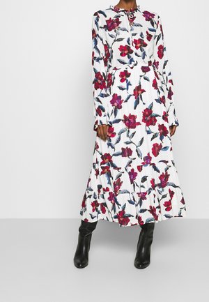 Kobieta w białej, długiej sukience w kwiaty z czerwonymi i fioletowymi kwiatami, z długimi rękawami i czarnymi botkami, stojąca na tle gładkiej ściany.