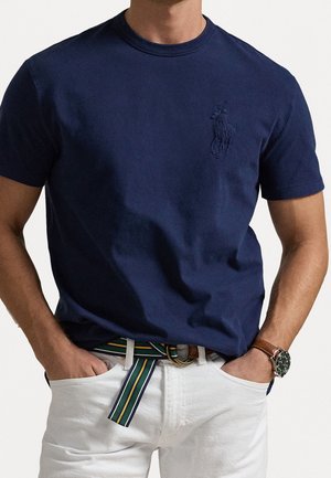 Mand iført navyblå t-shirt med lille broderet logo, hvide bukser, stribet stofbælte og armbåndsur med brun læderrem.