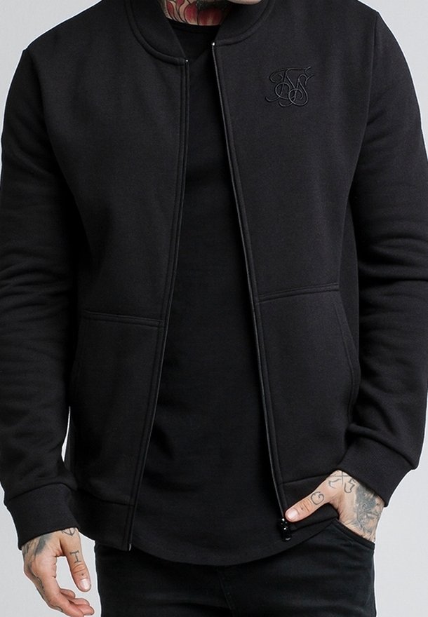 Homme portant une veste zippée noire unie avec un logo brodé discret sur la poitrine et un pantalon noir, mains partiellement visibles avec des tatouages.