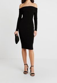 Žena v černých šatech s odhalenými rameny, sahajících po kolena, která drží černou kabelku na ruku a má na sobě černé lodičky na kotníkovém pásku.