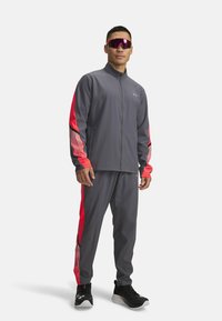 Gray athletic tracksuit with red side stripes; zip-up jacket, long sleeves; matching pants with striped accents; black sneakers and reflective sunglasses.