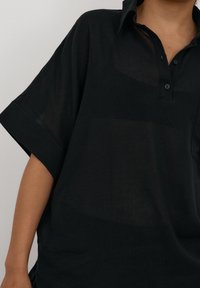 Czarna koszula z krótkim rękawem, wykonana z lekkiego materiału. Posiada kołnierzyk, listwę z guzikami oraz luźny fason z lekko przezroczystą fakturą.