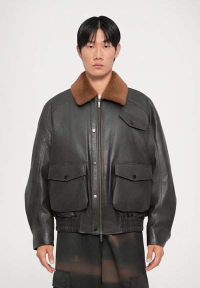 Veste bomber en cuir noir avec un col en fausse fourrure marron, comportant deux poches avant, une taille élastiquée et une fermeture éclair à l'avant.