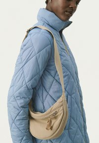 Borsa a tracolla beige con forma curva, tessuto texturizzato e chiusura con zippers. Indossata sopra un giubbotto trapuntato azzurro chiaro.