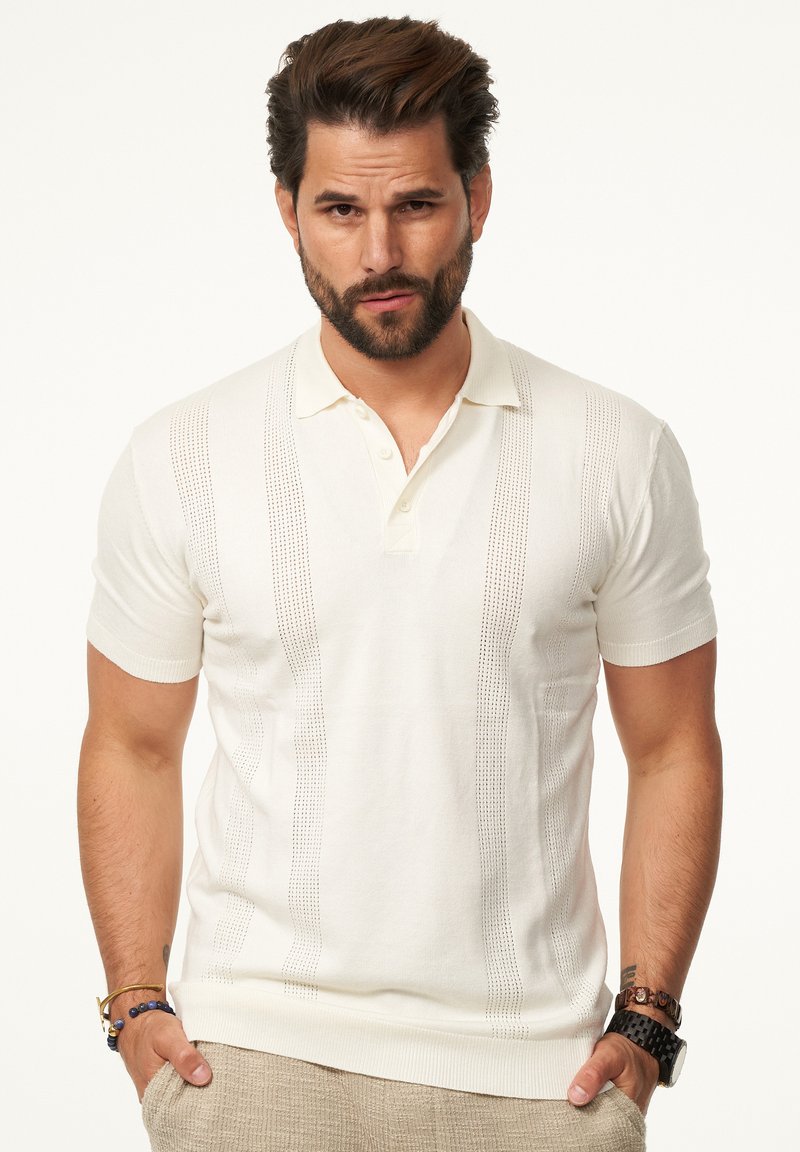 Weißes Polo-Shirt mit kurzen Ärmeln, gerippten Bündchen und vertikalen strukturierten Streifen. Kombiniert mit hellbeigen Hosen und Zubehör am Handgelenk.